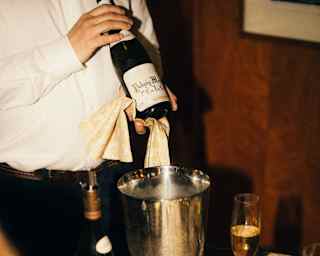 At a table laid for dining, with wineglasses and gold-plated cutlery, a waiter shows guests the label on a bottle of wine.