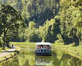 The Fleur de Lys cruises up the serene Ouche river, bordered by big trees and green banks, where a cyclist enjoys the route.
