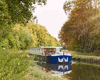 Front view of a luxurious river barge sailing down a sun-splashed tree-lined canal