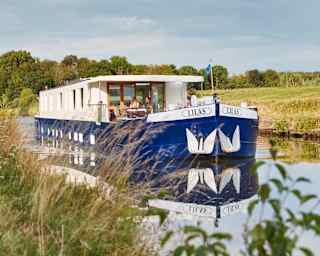 The Lilas cruising with two passengers on deck enjoying the sunshine and tranquillity, pictured through grasses on the bank.