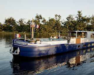 A fisherman stands on a lush grassy bank as the nose of a blue and white bateau edges around a waterway corner towards him.