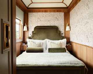 A cosy bedroom with a large plush olive-green headboard, matching pillows, white bedding, wood-panelled walls, patterned wallpaper, wall-mounted lamps, and a window letting in natural light.
