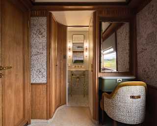 A luxurious train compartment with wood-panelled walls, patterned wallpaper, a dressing table with a mirror, an upholstered chair, and a view into a small bathroom with gold fittings and elegant lighting.
