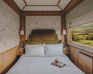 A cosy, neatly made bed in a vintage train compartment with elegant wood panelling, soft lighting, tea set on the bed, and a window showing a blurred green countryside landscape outside.