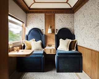 A cosy train compartment with two plush navy blue seats, patterned cushions, a small table set with drinks, wood panelling, and a window showing blurred scenery outside.