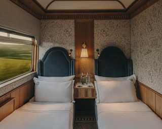 A cosy train compartment with two single beds, white linen, a wall lamp, and a small table with water glasses between the beds. The window shows a blurred green landscape passing by.