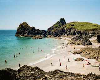 People relax and swim at a sandy beach surrounded by rocky cliffs and clear blue water under a sunny sky. Green hills rise in the background.