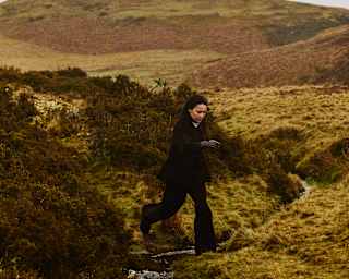 A person dressed in black jumps over a narrow stream in a grassy, hilly landscape under an overcast sky.