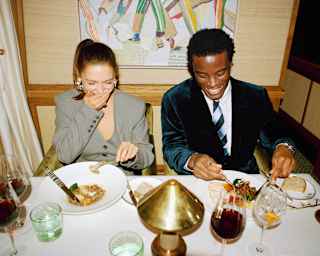 A woman and man dressed formally sit at a restaurant table, laughing and enjoying their meal. Wine glasses, plates with food, and a lamp are on the table, with colourful artwork hanging on the wall behind them.