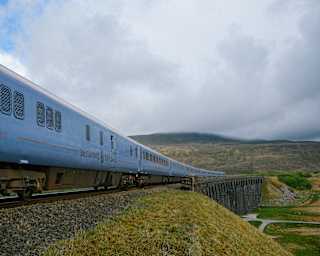 A blue passenger train travels across a bridge in a rural, hilly landscape with cloudy skies overhead.
