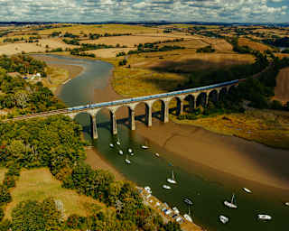 An aerial view of a train crossing a stone railway viaduct over a river with small boats moored in the water, surrounded by rolling hills and fields.