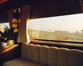 A cosy, upholstered bench sits by a large window on a train. Light curtains are gathered at the sides, and potted plants and lamps are on a nearby table. Outside, a sunlit rural landscape stretches into the distance.