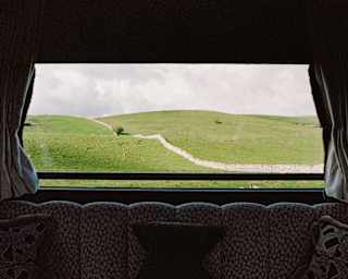 A green hillside with a rocky path is seen through a large window framed by dark curtains, behind a patterned sofa with cushions inside a cosy room.