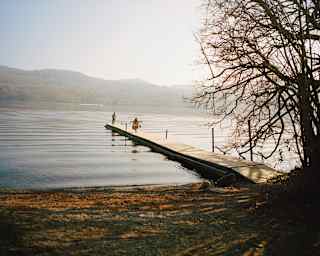 A wooden jetty extends into a calm lake, with two people standing near the end. Ripples spread across the water, and tree branches frame the right side. Distant hills are visible under a hazy sky.