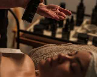 A person dispenses skincare product into their hand whilst another person lies on a massage table with a towel wrapped around their head, ready for a facial treatment. Bottles are visible in the background.