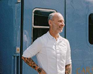 A man with tattoos wearing a white shirt stands and smiles in front of a blue train carriage on a sunny day.