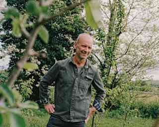 A smiling bald man in a denim jacket stands outdoors in a grassy area with trees and greenery in the background, partly framed by tree branches in the foreground.
