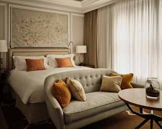 A taupe sofa with bronze cushions abuts a white bed with matching cushions, in a Deluxe Room with bright translucent drapes.