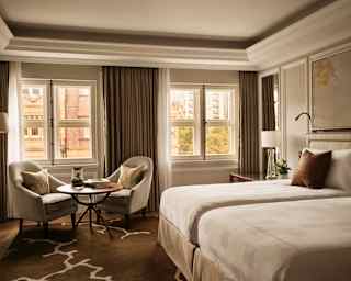View of a Junior Twin Suite in serene beige and brown décor, with white beds, two windows, and a table with a pair of chairs.