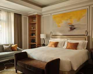 A yellow and grey abstract artwork hangs above a white Junior Suite bed with bronze cushions and a brown velvet bed bench.
