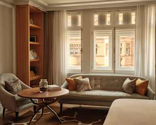 A three-pane window spills light onto a wide grey sofa in a Junior Suite living area with a lounge chair, table and bookcase.
