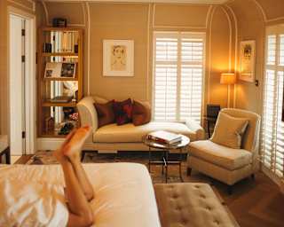 A guest's legs are seen on a bed in a view of mellow suite with backlit cornice, which glows above a chaise long and armchair.