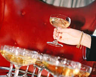 A hand lifts as glass of Champagne from a tray full of filled glasses, set on a red leather seat, seen in an angled close-up.