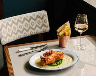 Close-up of crispy battered fish atop a bed of peas in a tartare-style sauce, served with a silver pot of chips and white wine.