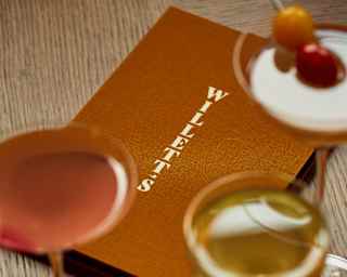 View of a burnt-orange menu wallet embossed with Willett's, written vertically in gold, seen over five drinks in soft-focus.
