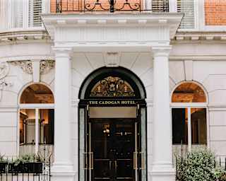 The Cadogan Hotel entrance with a grand white stone portico, flanked by arched windows which glow with interior lighting.