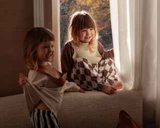 Two small girls, one sitting in a sunny window with half-drawn drapes, laugh as they enjoy time in their family suite.
