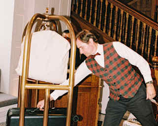 A hotel porter in a tartan waistcoat, white shirt and black trousers reaches for a case on a luggage trolley in the lobby.