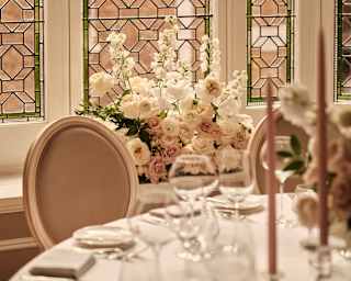 Geometric lead-pane windows spill light into a venue adorned with white and pink flowers, seen over a table in soft-focus.