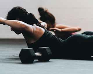 Next to a dumbbell, two women in black active wear exercise, lying on the floor on their stomachs with upper bodies raised.