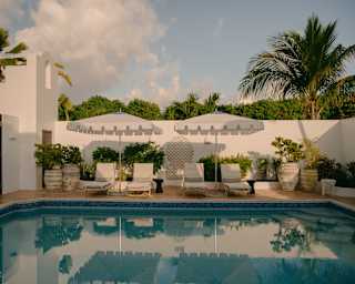 View over the still pool to two pairs of sun loungers, each sharing a white parasol, against a white wall with potted plants.