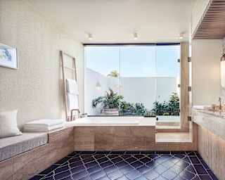 A spacious bathroom with black tiled floor, glass wall and large white tub features honey marble and cushioned bench seat