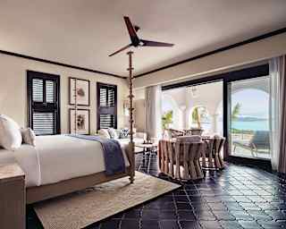 Dark wood plantation shutters give privacy to an elegant bedroom with crisp white four poster, sitting area and sea views