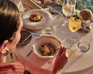 A couple share a meal with white wine and a cocktail at a Pimms terrace table overlooking the ocean, seen from a high angle.