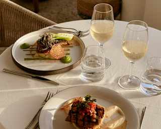 A round table set for two with elegant crockery, glasses of white wine, water, and neatly arranged cutlery. Plates hold gourmet meals with sauce and vegetables, all in natural sunlight.