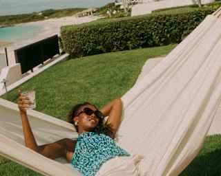A woman in shades and patterned blue top lies back in a white hammock with a drink, in a view over a private villa garden.