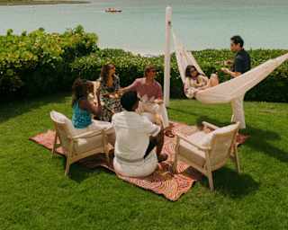 Guests chat in a lush green sea-view garden, with two on chairs, two on a blanket, one in a hammock and another standing.