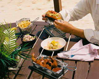 Looking across a vibrant Cap Shack lunch, enjoyed by a guest at a wooden table, to the dazzling white beach and sapphire sea.