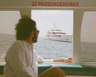 On the ferry to Anguilla, a dark-haired man with sunglasses and cool white shirt gazes out of the window at a nearby yacht.