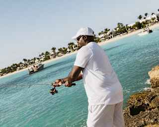 A man dressed in white shorts, shirt and cap casts his fishing rod from the rocks on the east headland of Maundays Bay.