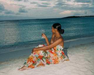 A woman in a bright orange-hued floral dress sits on the white sand by the calm evening ocean with a drink, seen from behind.