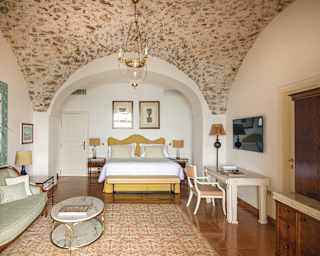 Polished russet floor tiles reflect a butter yellow vallance, crisp white linens top a king bed near a gilt edged white chair