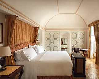 A king bed with white linens and heavy silk amber drapes sits centre of a Romanesque-styled room with ochre wall edge lines