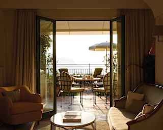 Hotel room lounge area with open patio doors and a sunlit balcony beyond