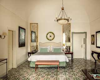 A bed’s white linens and apple green valance and pillows, echoes the greens in the multicoloured hand painted floor tiles