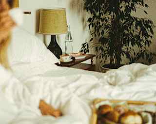 Soft-focus image of a woman sitting in bed beneath a white duvet, sipping tea as she enjoys breakfast from a tray on her lap.
